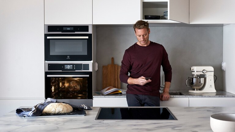 Homme dans une cuisine utilisant son téléphone portable derrière un comptoir où se trouvent une plaque de cuisson et du pain