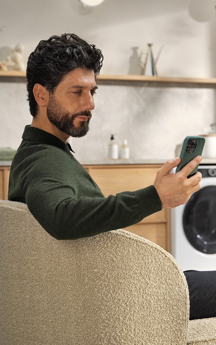 Un homme assis sur une chaise avec un téléphone à la main et une machine à laver en arrière-plan.